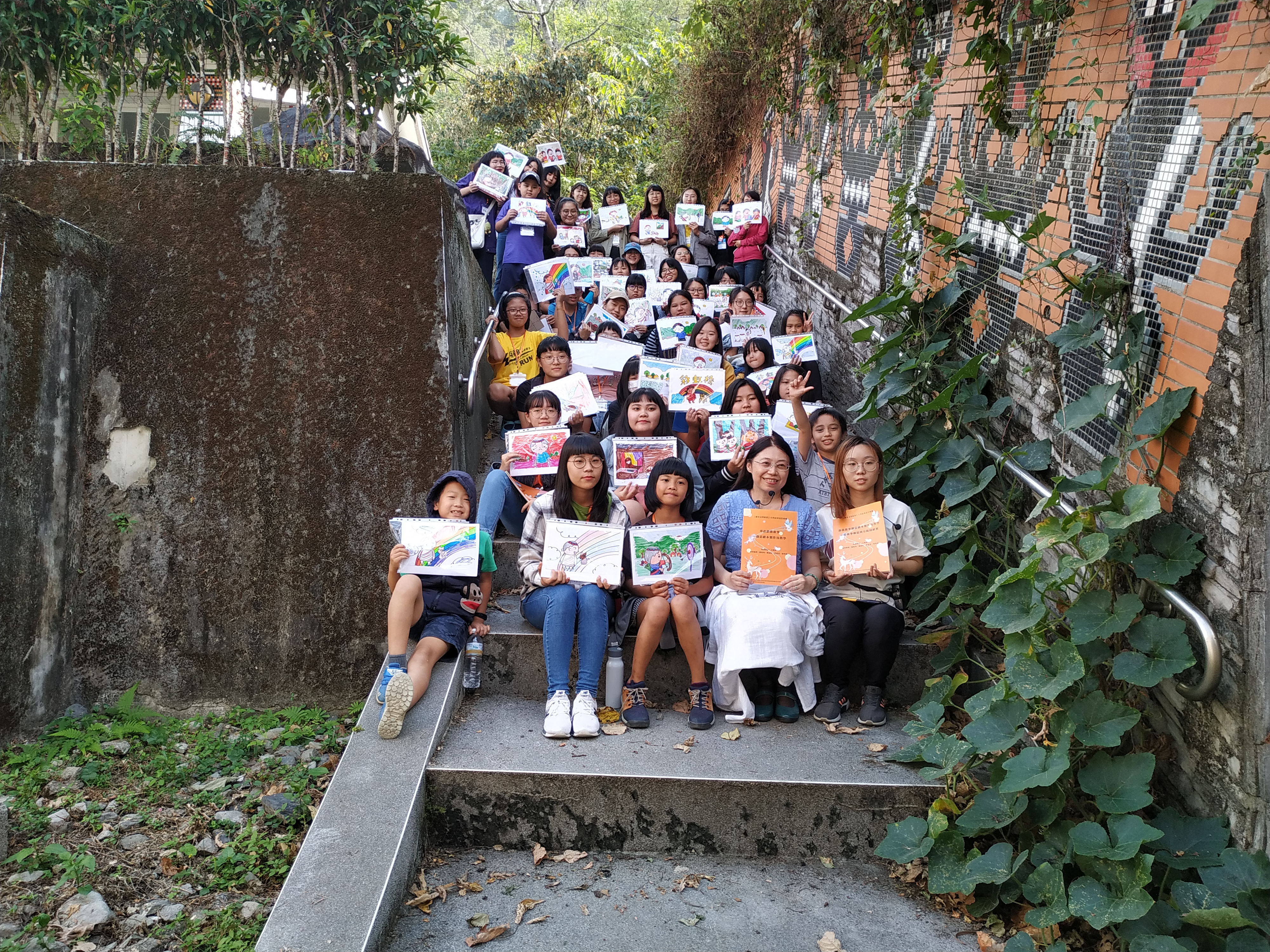 王晴慧老師(前排右二)與亞大學生、松鶴部落博愛國小學生，手持共同完成的繪本分鏡圖，於博愛國小部落圖騰階梯旁大合照。